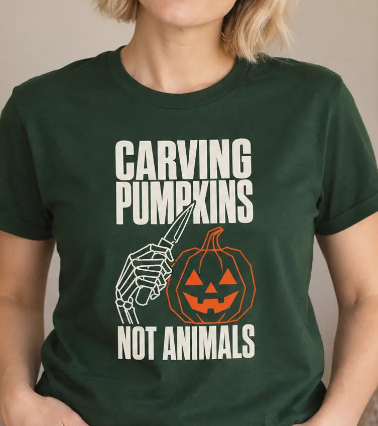 A woman wearing a dark green graphic t-shirt with the text 'CARVING PUMPKINS NOT ANIMALS' and a skeleton hand carving a pumpkin illustration, posed in a cozy indoor setting.