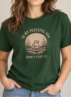 A woman wearing a dark green graphic t-shirt with a vintage-style frog illustration and the text 'IN MY PEACEFUL ERA (DON'T TEST IT)'.