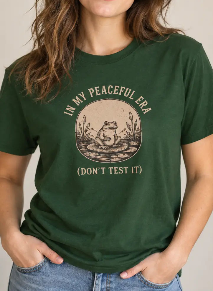 A woman wearing a dark green graphic t-shirt with a vintage-style frog illustration and the text 'IN MY PEACEFUL ERA (DON'T TEST IT)'.