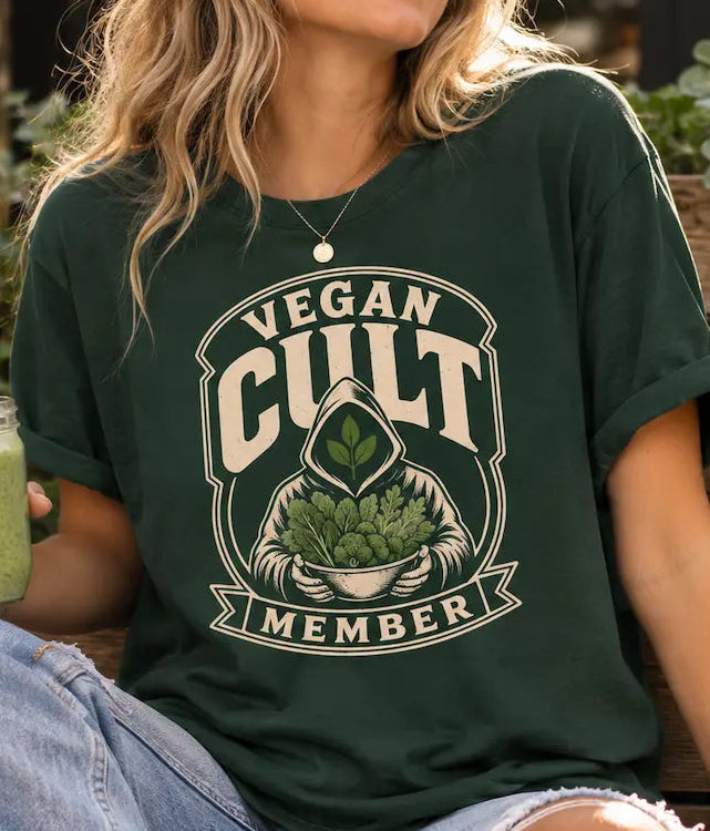 A woman wearing a dark green graphic t-shirt with a cream-colored 'Vegan Cult Member' design, sitting outdoors on a wooden bench and holding a green smoothie.