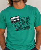 A man wearing a teal green graphic t-shirt with a black cassette tape and the text 'OUT OF SYNC WITH THE MAINSTREAM' printed on the front, set against a neutral textured wall.