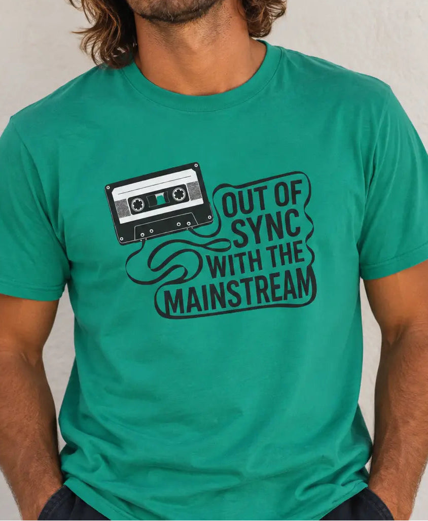 A man wearing a teal green graphic t-shirt with a black cassette tape and the text 'OUT OF SYNC WITH THE MAINSTREAM' printed on the front, set against a neutral textured wall.