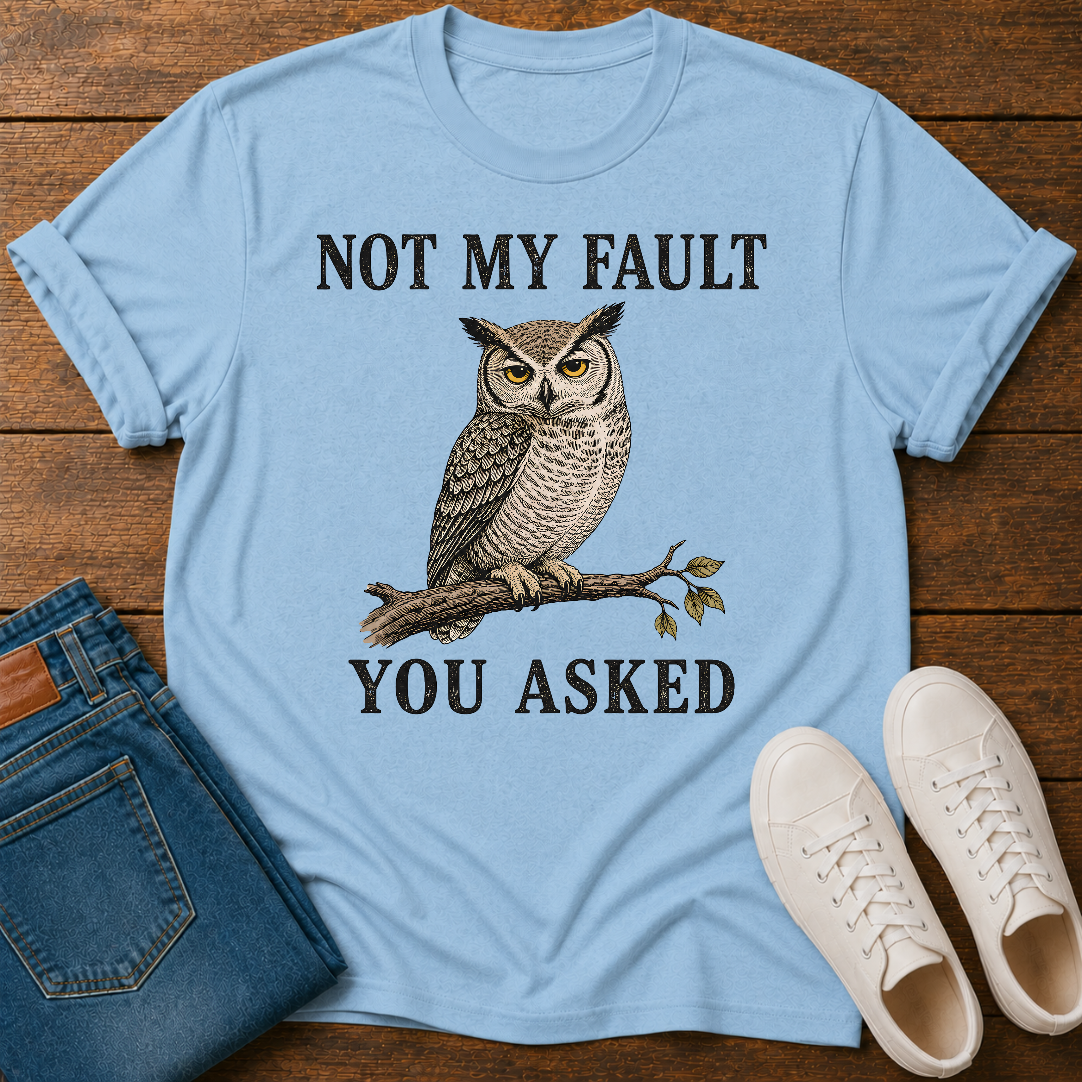 A light blue graphic t-shirt laid flat on a wooden surface, styled with blue jeans, white sneakers. The t-shirt features a graphic of a owl perched on a branch with the text 'NOT MY FAULT YOU ASKED' in black.