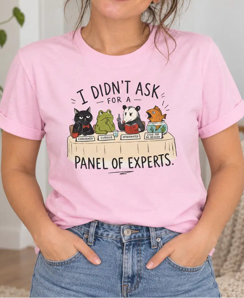 A woman wearing a light pink graphic t-shirt with a cartoon panel of animals and the text 'I DIDN'T ASK FOR A PANEL OF EXPERTS.' She is posing in a cozy, modern living room setting.