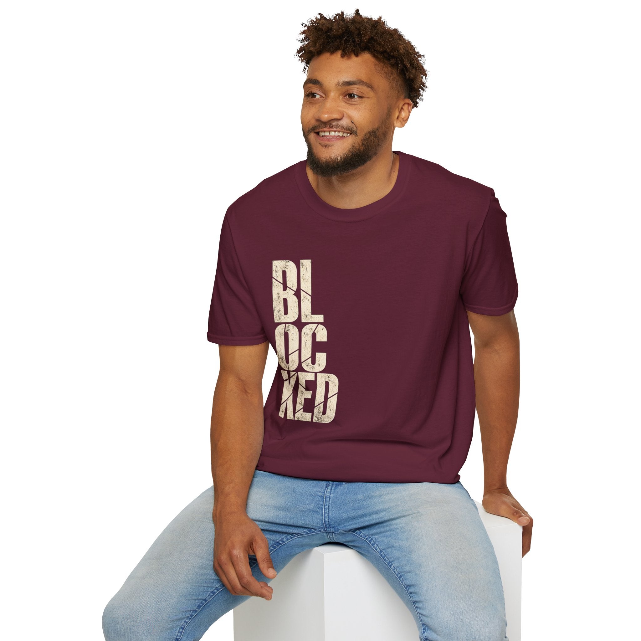 A man wearing a maroon crewneck t-shirt with the word 'BLOCKED' printed vertically in distressed beige letters, paired with light blue jeans, sitting on a white block against a plain white background.