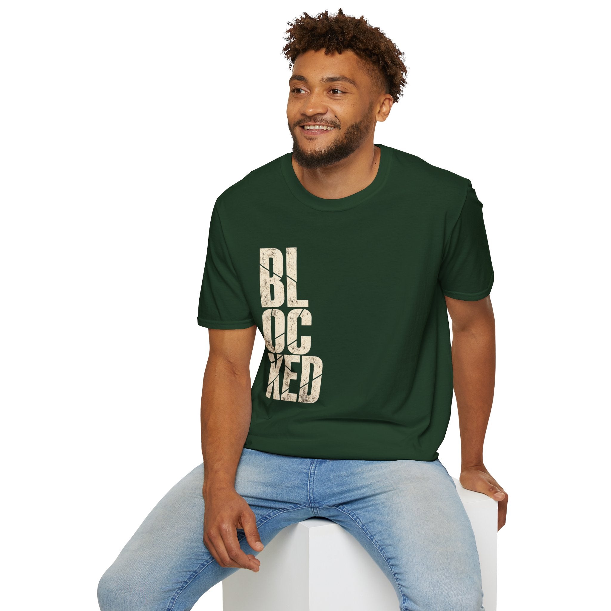 A man wearing a dark green crewneck t-shirt with the word 'BLOCKED' printed vertically in distressed beige block letters, paired with light blue jeans, sitting on a white block against a solid white background.