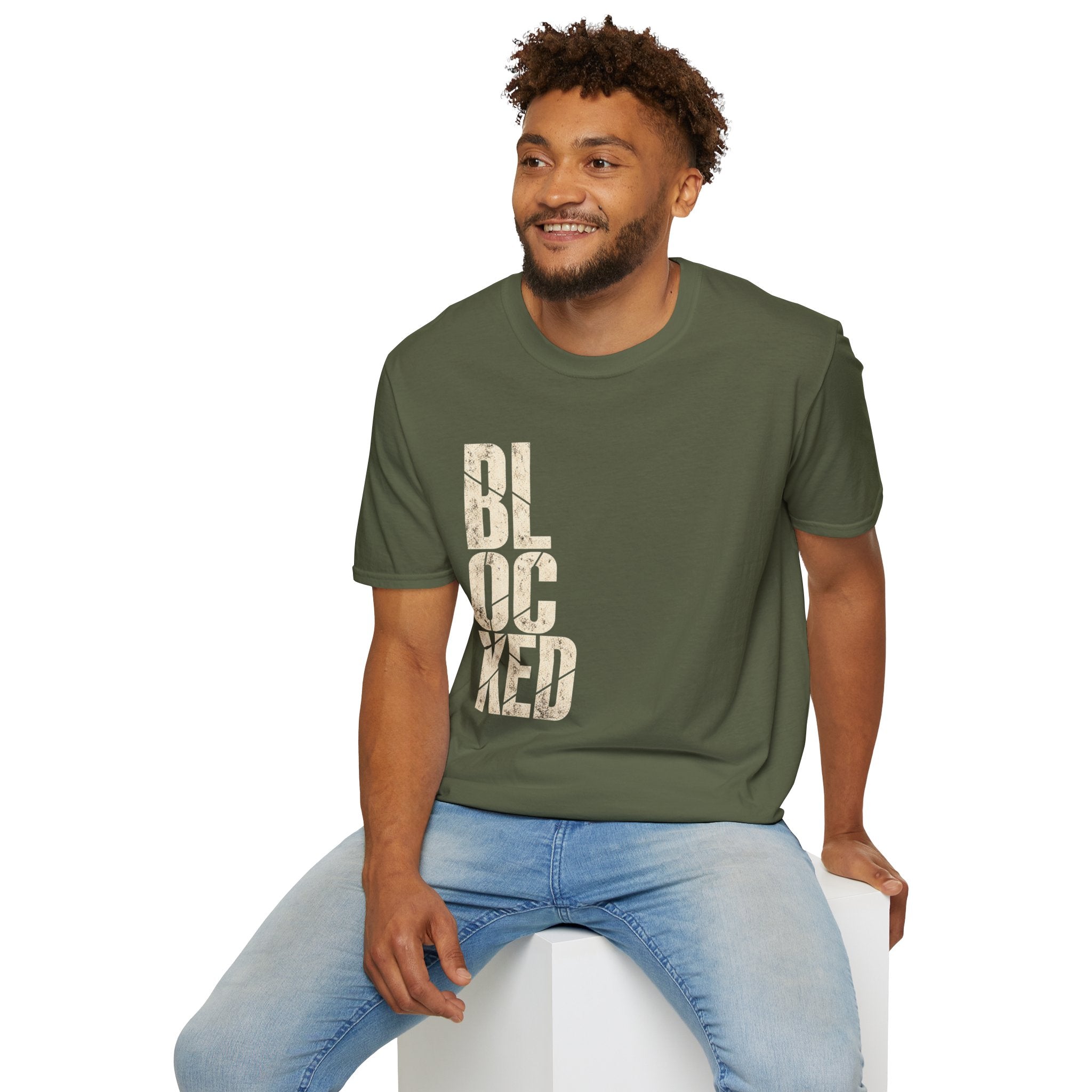 A man wearing an military green t-shirt with a distressed vertical graphic that reads 'BLOCKED' in a light beige color, paired with light blue jeans, sitting on a white block against a plain white background.