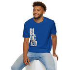 A man wearing a royal blue crewneck t-shirt with the word 'BLOCKED' printed vertically in distressed white letters, paired with light-wash blue jeans, sitting on a white block against a plain white background.
