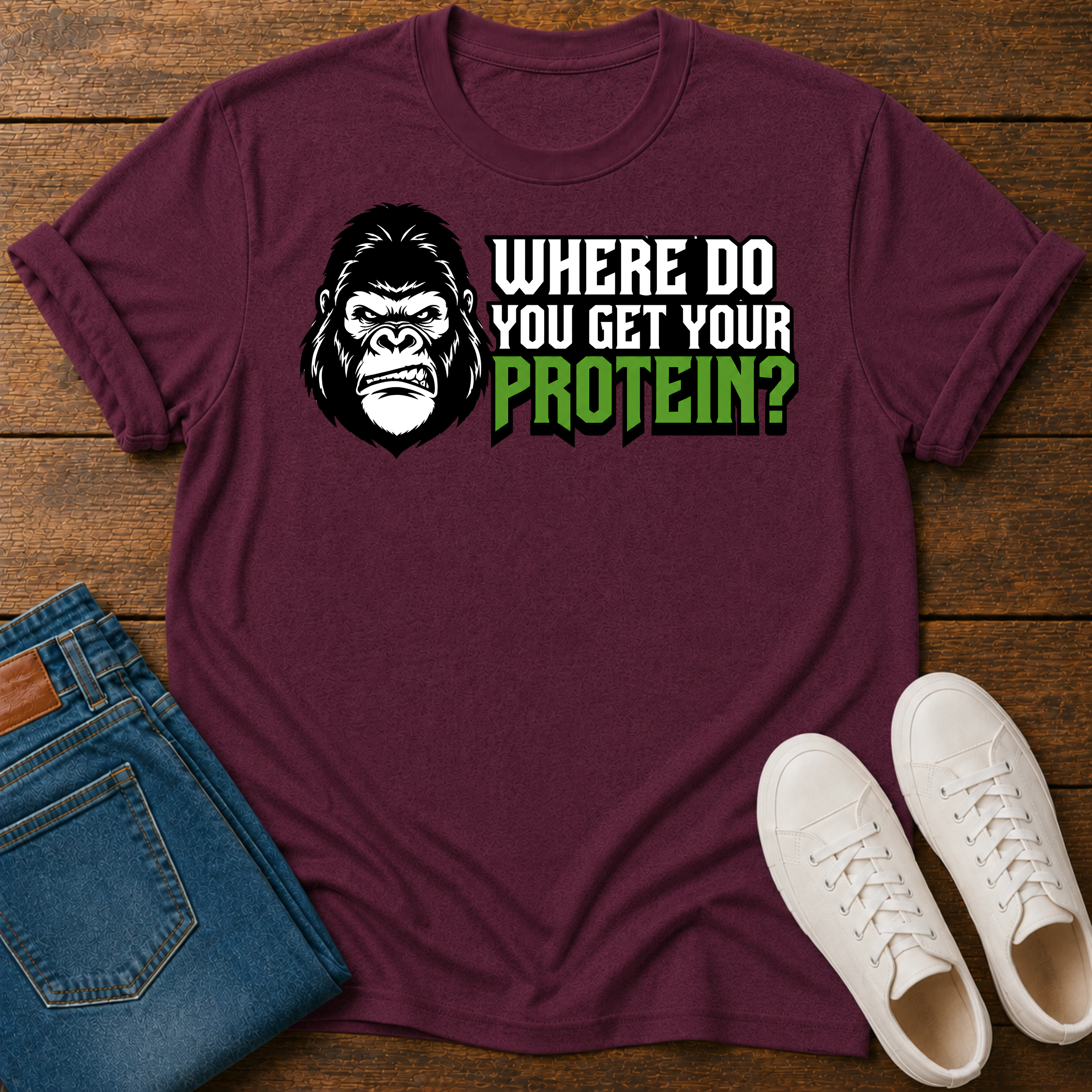 A maroon short-sleeve graphic T-shirt laid flat on a wooden surface, styled with folded blue jeans and white sneakers. The shirt features a large gorilla skull design with leaf details, crossed plant stems, and the text ''where do you get your protein from?"