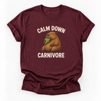 A maroon short-sleeve t-shirt with a graphic of a capybara eating green leaves, surrounded by the text 'CALM DOWN CARNIVORE' in cream lettering, displayed on a clean white background.