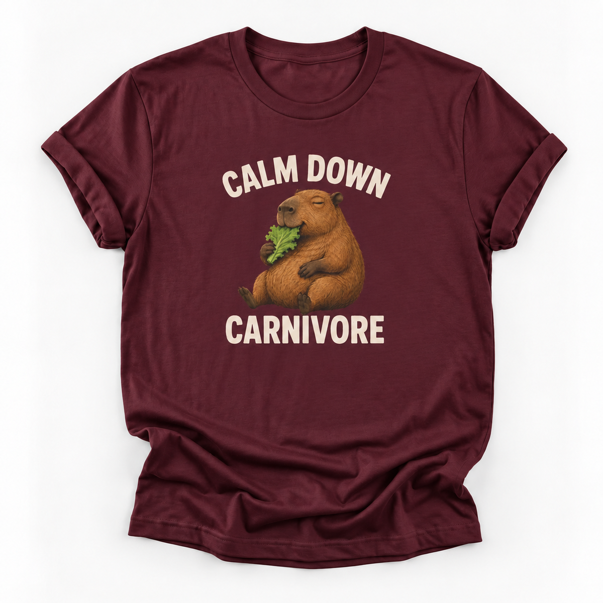 A maroon short-sleeve t-shirt with a graphic of a capybara eating green leaves, surrounded by the text 'CALM DOWN CARNIVORE' in cream lettering, displayed on a clean white background.