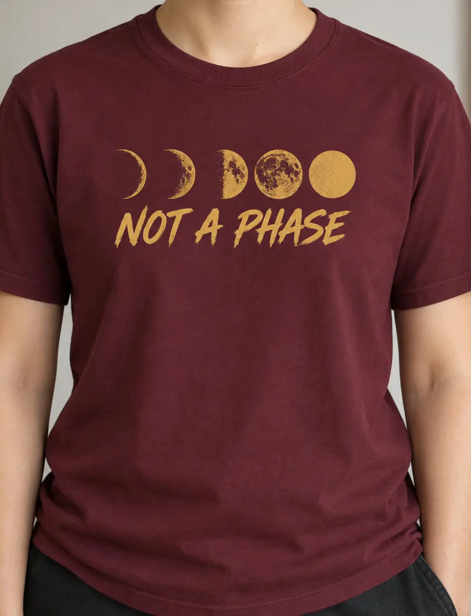 A person wearing a maroon short-sleeve t-shirt with a gold moon phase graphic and the text 'NOT A PHASE' printed on the chest, shown in an indoor setting.