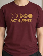 A person wearing a maroon short-sleeve t-shirt with a gold moon phase graphic and the text 'NOT A PHASE' printed on the chest, shown in an indoor setting.