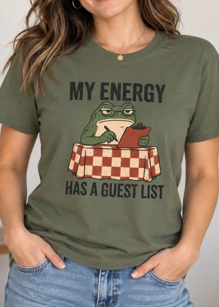 A woman wearing an olive green graphic t-shirt with a cartoon frog sitting at a checkered table reading a book, with the text 'MY ENERGY HAS A GUEST LIST'.