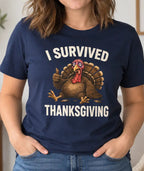 A woman wearing a navy blue graphic t-shirt with a turkey illustration and the text 'I SURVIVED THANKSGIVING' in white, standing in a cozy living room setting.