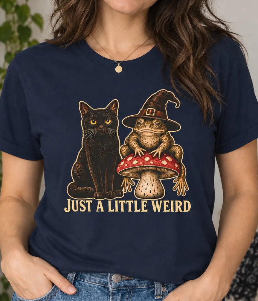 A woman wearing a navy blue graphic t-shirt with a design of a black cat and a toad wearing a witch hat on a mushroom, with the text 'JUST A LITTLE WEIRD'.