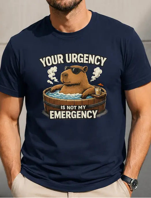 A man wearing a navy blue graphic t-shirt with a cartoon capybara in a hot tub and the text 'YOUR URGENCY IS NOT MY EMERGENCY'. He is wearing beige pants and sunglasses, standing against a concrete wall.