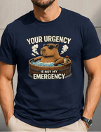 A man wearing a navy blue graphic t-shirt with a cartoon capybara in a hot tub and the text 'YOUR URGENCY IS NOT MY EMERGENCY'. He is wearing beige pants and sunglasses, standing against a concrete wall.
