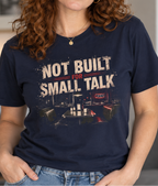 A woman with curly red hair wearing a navy blue graphic t-shirt that reads 'NOT BUILT FOR SMALL TALK' in distressed white and red lettering, paired with blue jeans, set against a softly blurred indoor background.