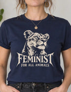 A woman wearing a navy blue graphic t-shirt with a white illustration of a lion and the text 'FEMINIST FOR ALL ANIMALS'. She is standing in a brightly lit room with plants and home decor in the background.