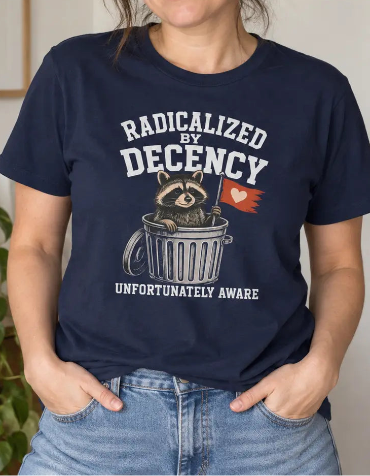 A woman wearing a navy blue graphic t-shirt with a cartoon sloth in a trash can and the text 'RADICALIZED BY DECENCY UNFORTUNATELY AWARE'. She is standing in a brightly lit room with a wooden dresser, a green plant, and a framed print on the wall.
