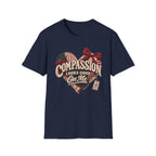 A navy blue short-sleeve crewneck t-shirt featuring a distressed graphic on the front. The graphic shows a heart made of red and white gingham patterns, with a red bow on top, and the text 'COMPASSION LOOKS GOOD On Me' in a rustic cream font.
