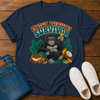 A navy blue graphic t-shirt featuring a gorilla sitting with bananas, peanuts, and a coconut under the text 'PROTEIN DEFICITY SURVIVOR'. The shirt is styled in a flat-lay on a wooden background alongside folded blue jeans and a pair of white sneakers.