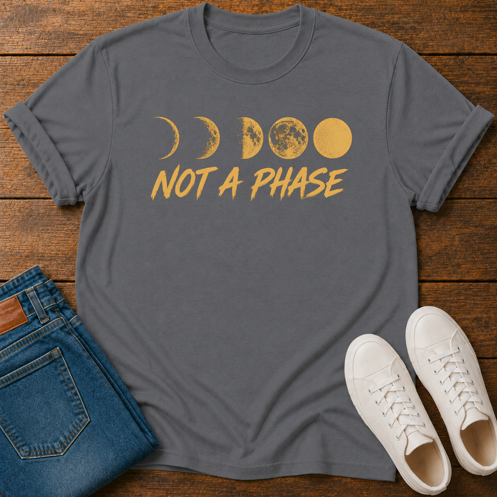 A dark gray crewneck t-shirt with a yellow moon phase graphic and the text 'NOT A PHASE' printed on the front. The shirt is styled in a flat-lay arrangement on a wooden surface, accompanied by folded blue jeans and a pair of white sneakers.