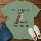 A sage graphic t-shirt laid flat on a wooden surface, styled with blue jeans, white sneakers. The t-shirt features a graphic of a owl perched on a branch with the text 'NOT MY FAULT YOU ASKED' in black.