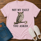 A pink graphic t-shirt laid flat on a wooden surface, styled with blue jeans, white sneakers. The t-shirt features a graphic of a owl perched on a branch with the text 'NOT MY FAULT YOU ASKED' in black.