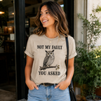 A woman wearing a beige graphic t-shirt with a black owl illustration and the text 'NOT MY FAULT YOU ASKED', paired with blue jeans and a black shoulder bag, standing in front of a storefront.