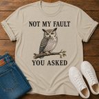 A beige graphic t-shirt laid flat on a wooden surface, styled with blue jeans, white sneakers. The t-shirt features a graphic of a owl perched on a branch with the text 'NOT MY FAULT YOU ASKED' in black.
