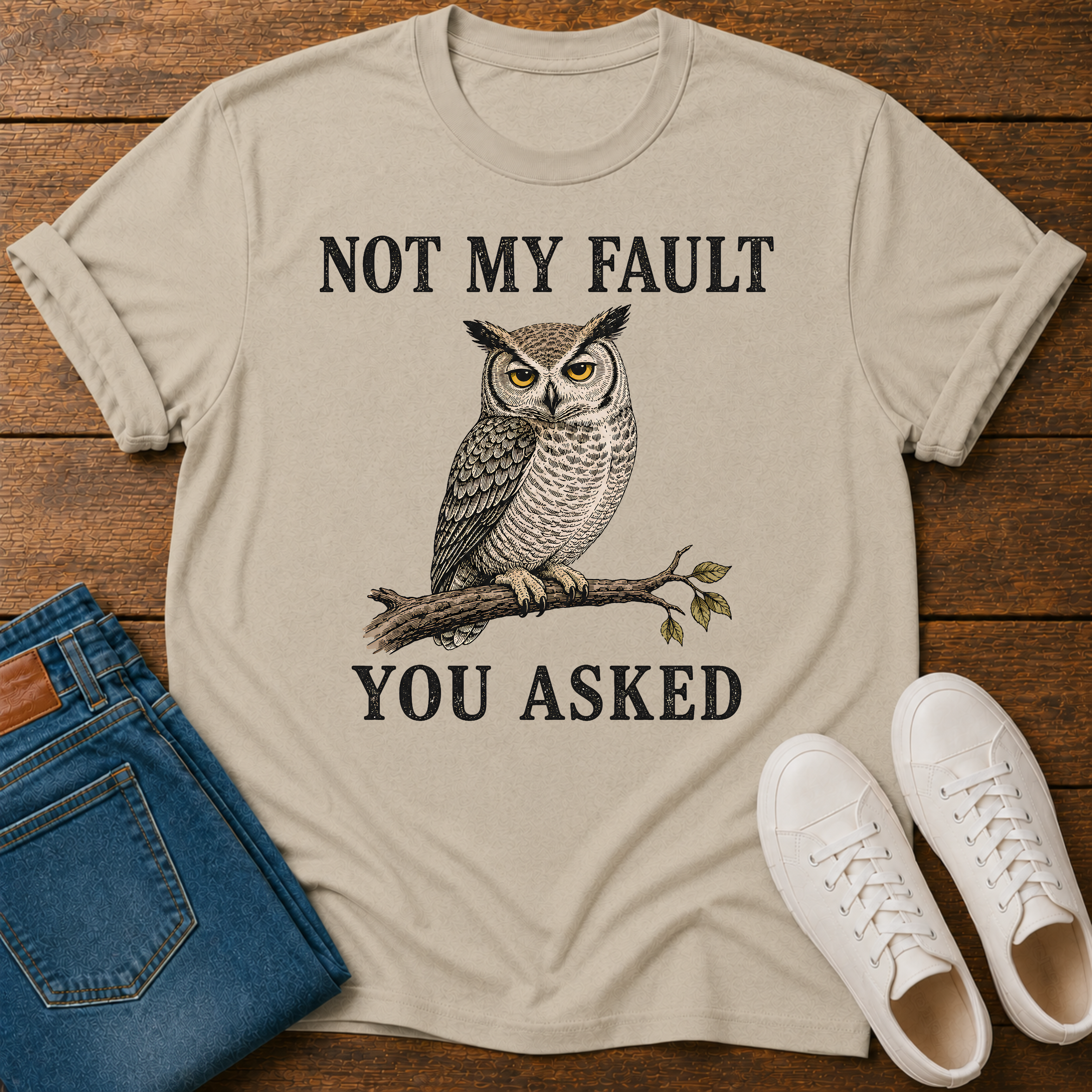 A beige graphic t-shirt laid flat on a wooden surface, styled with blue jeans, white sneakers. The t-shirt features a graphic of a owl perched on a branch with the text 'NOT MY FAULT YOU ASKED' in black.