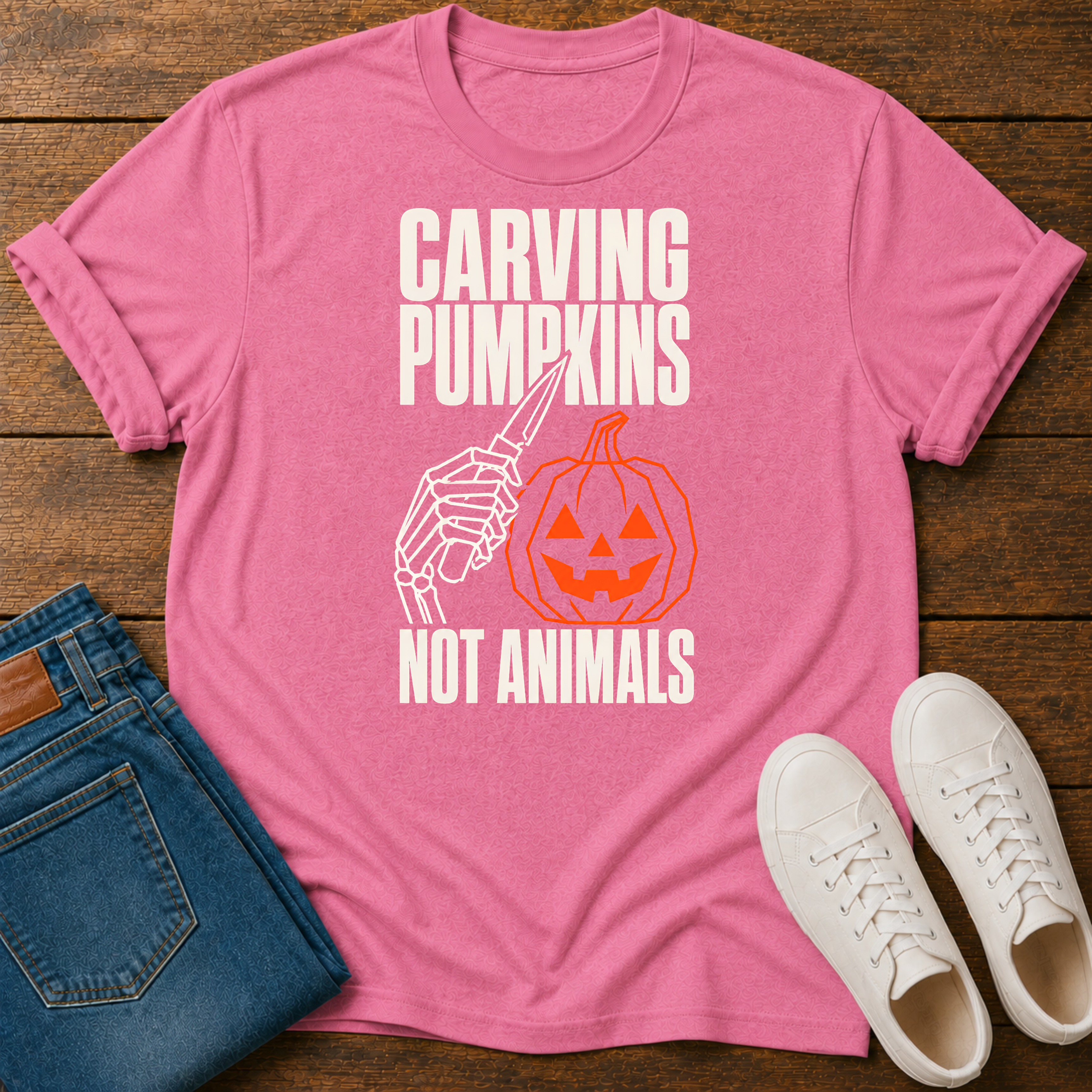 A pink short-sleeve graphic T-shirt laid flat on a wooden surface, styled with folded blue jeans and white sneakers. The shirt has a white and orange Halloween-themed graphic reading 'CARVING PUMPKINS NOT ANIMALS' with a skeleton hand holding a carving knife and a jack-o'-lantern.