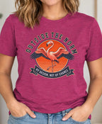 A woman wearing a heather magenta graphic t-shirt with a flamingo design and the text 'OUTSIDE THE NORM BY DESIGN, NOT BY CHANCE', paired with blue jeans in a brightly lit indoor setting.