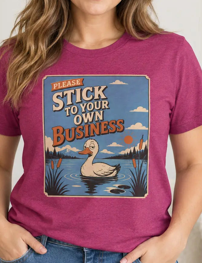 A woman wearing a heathered magenta graphic t-shirt with a duck pond design and the text 'PLEASE STICK TO YOUR OWN BUSINESS'.