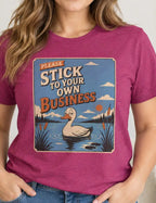 A woman wearing a heathered magenta graphic t-shirt with a duck pond design and the text 'PLEASE STICK TO YOUR OWN BUSINESS'.