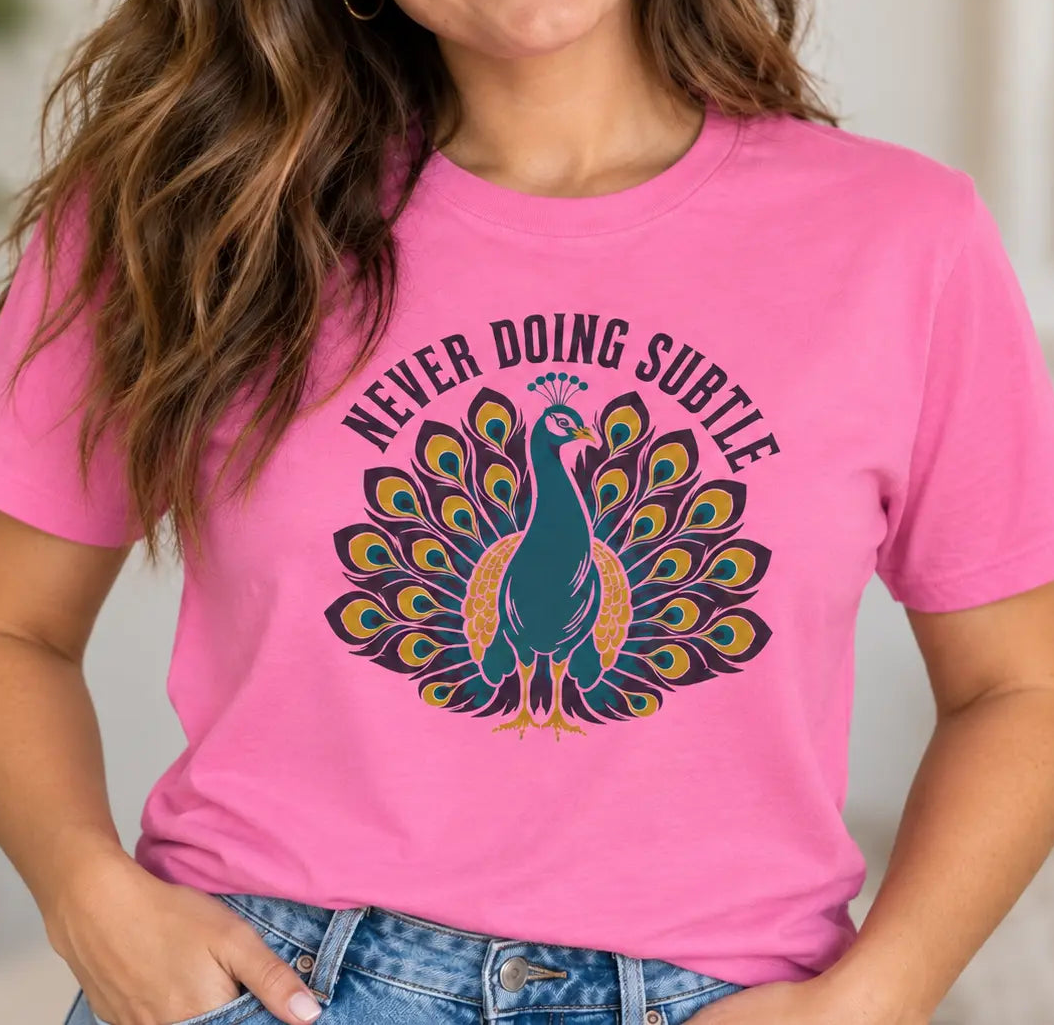 A woman wearing a bright pink graphic t-shirt with a colorful peacock design and the text 'NEVER DOING SUBTLE'.