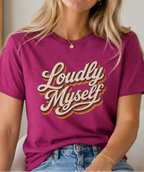 A woman wearing a magenta short-sleeve graphic t-shirt with the words 'Loudly Myself' in a cream and gold retro script, paired with blue jeans, sitting in a brightly lit indoor setting.