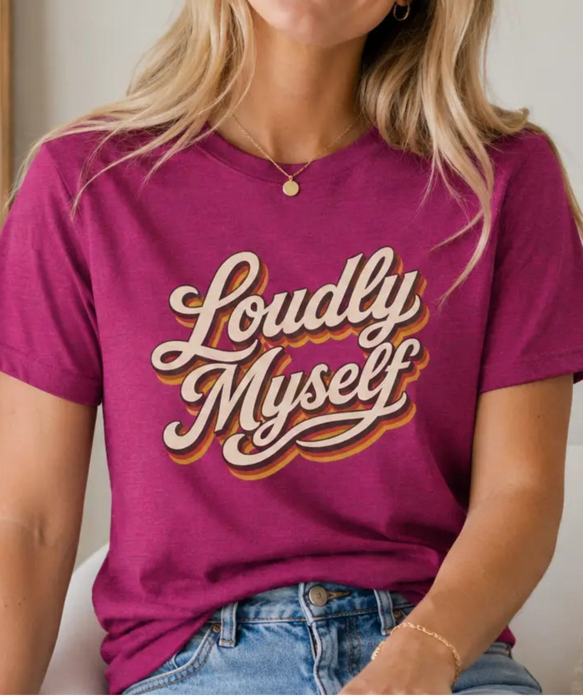 A woman wearing a magenta short-sleeve graphic t-shirt with the words 'Loudly Myself' in a cream and gold retro script, paired with blue jeans, sitting in a brightly lit indoor setting.