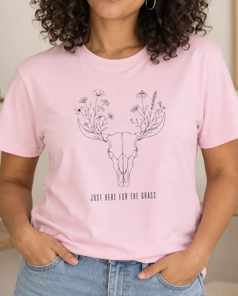 A woman with curly dark hair wearing a light pink vegan graphic t-shirt with a black line art deer skull and floral design, with text 'just here for the grass' paired with blue jeans in a cozy, sunlit room.