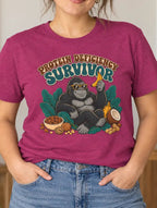 A woman wearing a heather magenta graphic t-shirt with a gorilla and 'PROTEIN DEFICIENCY SURVIVOR' print, paired with blue jeans in a cozy, plant-filled room.