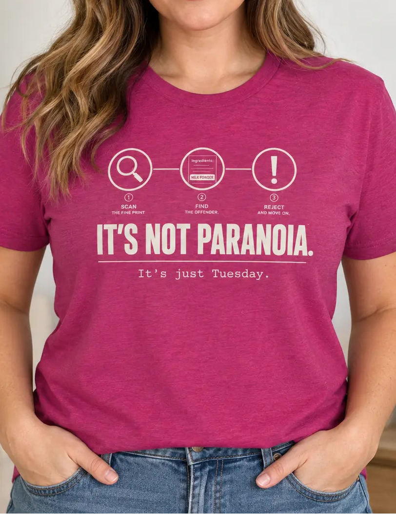 A woman wearing a heather magenta graphic t-shirt with white text that reads 'IT'S NOT PARANOIA. It's just Tuesday.