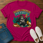 A bright pink graphic t-shirt laid flat on a wooden surface, styled with folded blue jeans and white sneakers. The t-shirt features a cartoon gorilla wearing sunglasses and holding a banana, surrounded by leaves and snacks, with the text 'PROTEIN DEFICITY SURVIVOR' above it.