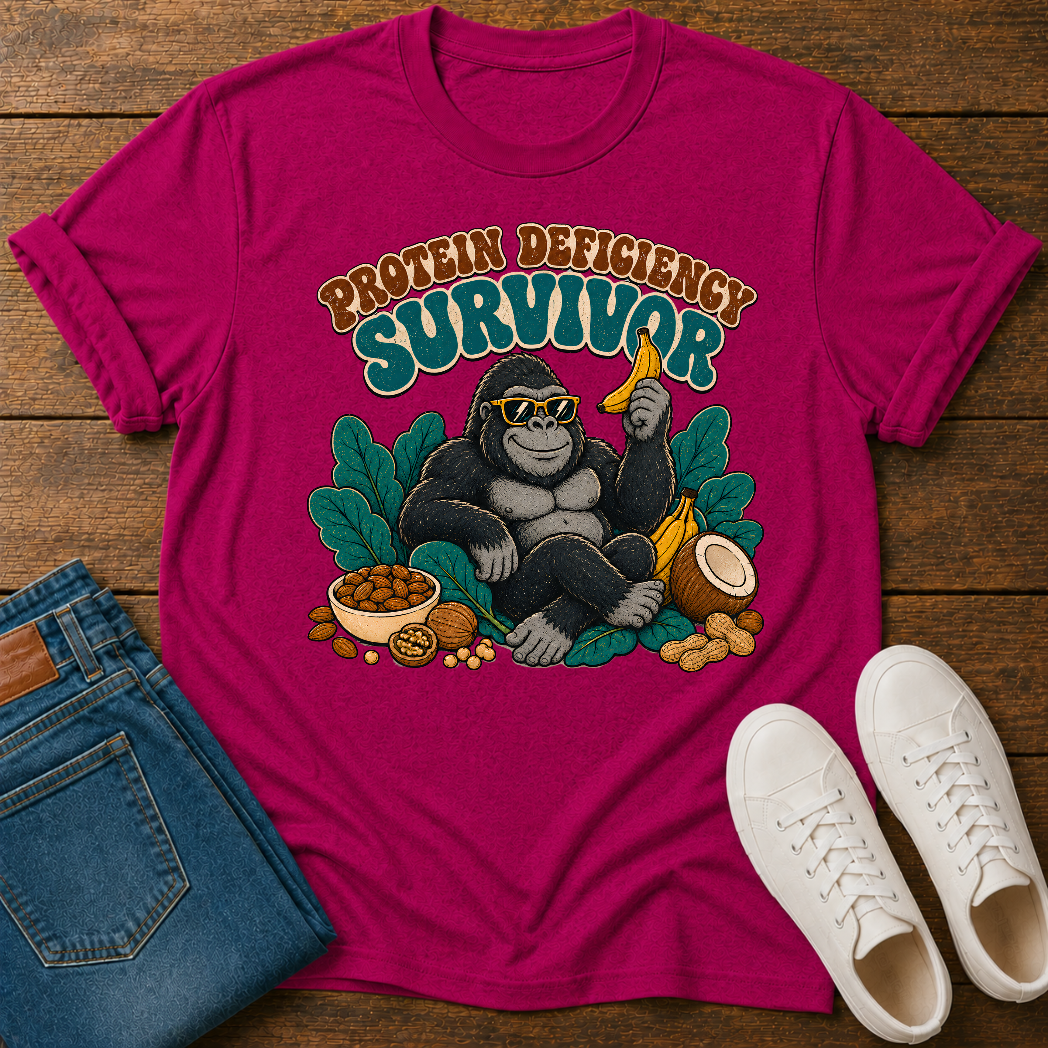 A bright pink graphic t-shirt laid flat on a wooden surface, styled with folded blue jeans and white sneakers. The t-shirt features a cartoon gorilla wearing sunglasses and holding a banana, surrounded by leaves and snacks, with the text 'PROTEIN DEFICITY SURVIVOR' above it.