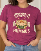 A woman wearing a heathered magenta graphic t-shirt with a cartoon hummus bowl and the text 'EMOTIONALLY SUPPORTED BY HUMMUS'. She is standing in a cozy, modern living room setting.