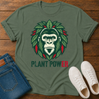 A military green graphic t-shirt laid flat on a wooden surface, styled with folded blue jeans and white sneakers. The t-shirt features a graphic of a gorilla face surrounded by green leaves with red accents, and the text 'PLANT POWER' printed below.