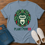 A stone blue crewneck t-shirt laid flat on a wooden surface, featuring a graphic of a gorilla face surrounded by green leaves with red accents, and the text 'PLANT POWER' below. The shirt is styled with folded blue jeans and a pair of white sneakers.