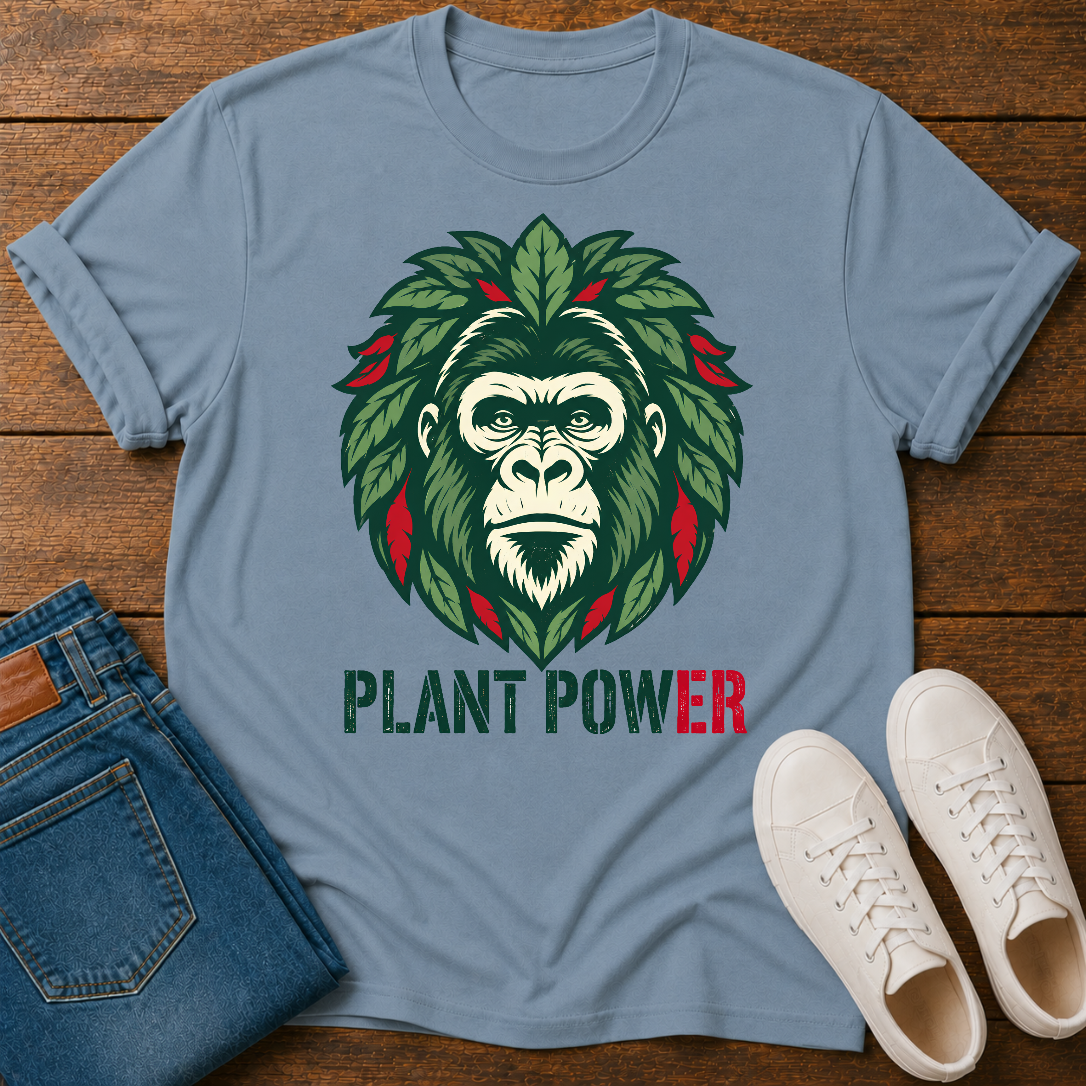 A stone blue crewneck t-shirt laid flat on a wooden surface, featuring a graphic of a gorilla face surrounded by green leaves with red accents, and the text 'PLANT POWER' below. The shirt is styled with folded blue jeans and a pair of white sneakers.