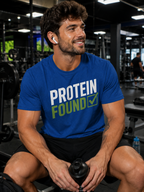 A man in a gym wearing a blue t-shirt with the text 'PROTEIN FOUND' in white and green, holding a black shaker bottle.